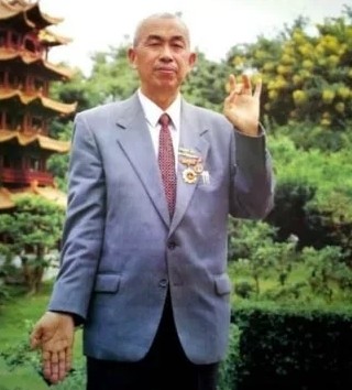 Tian Ruisheng in a grey suit with medals on his chest, one hand raised, in a garden with a pagoda behind him